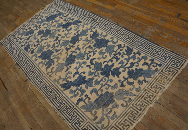 Blue and tan rug featuring large floral vines and a geometric fret border is displayed on distressed wood flooring.