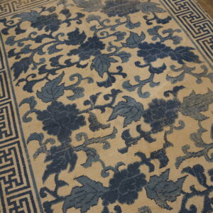 A blue and beige low-pile rug featuring a dense floral pattern and a Greek key border rests on wood flooring.