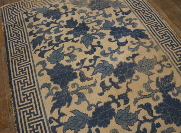 A blue and beige low-pile rug featuring a dense floral pattern and a greek key border rests on wood flooring.
