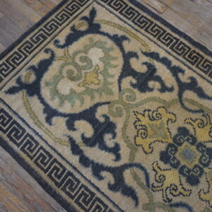 Tan rug with dark blue floral motifs and a Greek key border sits on rustic wood planks.
