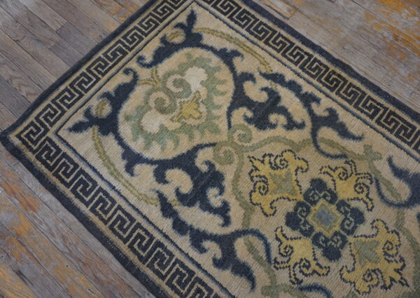 Tan rug with dark blue floral motifs and a greek key border sits on rustic wood planks.
