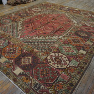 Low-pile geometric rug with complex red, green, orange, and brown patterns rests on worn wood planks.