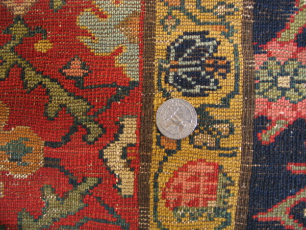 Close-up of a low-pile knotted rug showing red, gold, and navy geometric patterns next to a quarter coin.