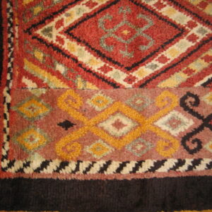 High-pile rug with geometric patterns in red, gold, and dark brown, lying on a wooden floor.