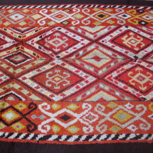 Textured rug featuring geometric diamond and scroll patterns in red, orange, yellow, and white, framed by dark brown borders.