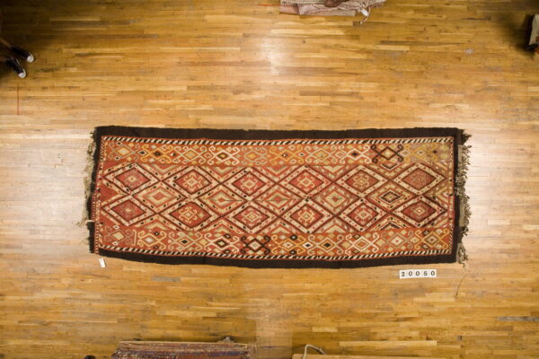 Rectangular rug with red, gold, and cream diamond patterns and a dark brown border on light wood flooring.