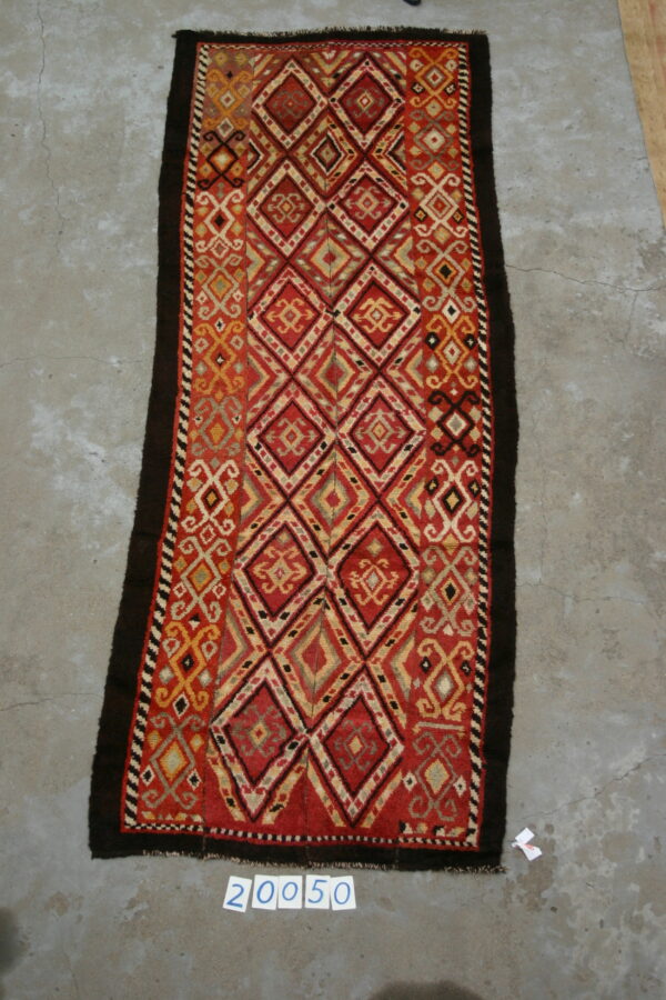Runner rug displaying a bright red and gold repeating diamond pattern with dark brown outer edges on concrete.