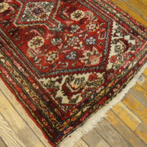 A low-pile red rug with cream and dark floral patterns rests diagonally on distressed, light wooden floorboards.