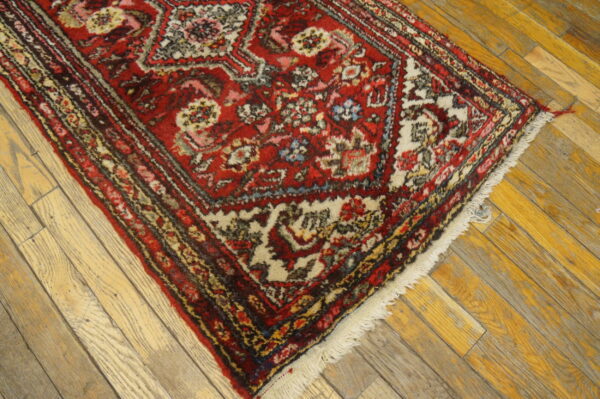 A low-pile red rug with cream and dark floral patterns rests diagonally on distressed, light wooden floorboards.