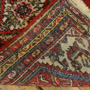 Red, yellow, and blue patterned rug corner folded over, showing the woven back structure on wood flooring.