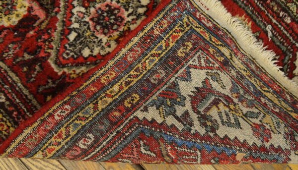 Red, yellow, and blue patterned rug corner folded over, showing the woven back structure on wood flooring.