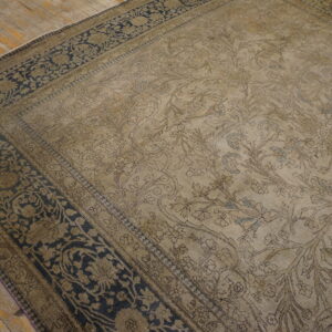 A muted tan rug with a floral and bird design and dark navy border rests on rustic wooden flooring.