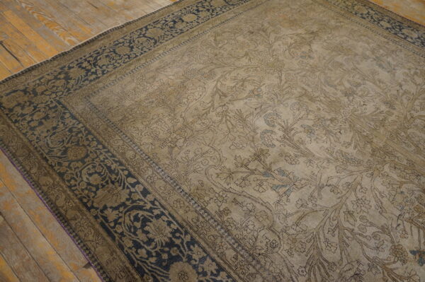 A muted tan rug with a floral and bird design and dark navy border rests on rustic wooden flooring.