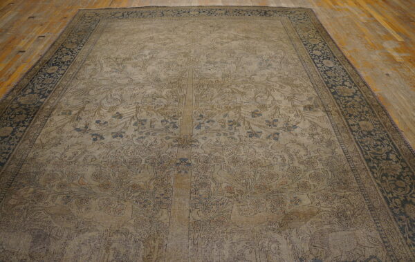 Faded tan rug with intricate tree of life pattern and dark blue border lying on distressed wood flooring.