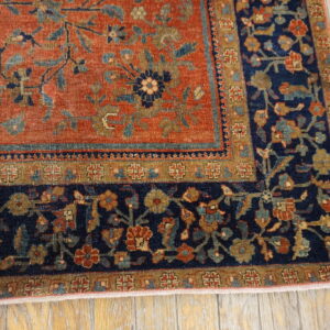 Close-up of a rug corner featuring a rusty orange field and a navy floral border on distressed wood flooring.