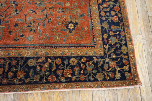 Close-up of a rug corner featuring a rusty orange field and a navy floral border on distressed wood flooring.