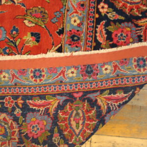 Close-up of a red and navy short-pile floral rug folded back, exposing the backing over wood plank flooring.
