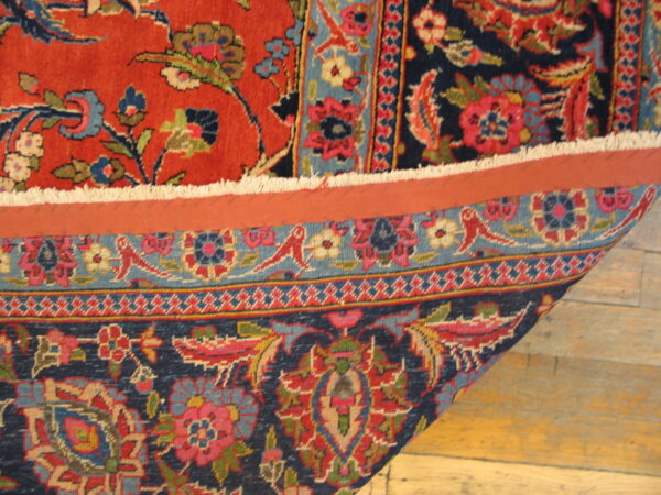 Close-up of a red and navy short-pile floral rug folded back, exposing the backing over wood plank flooring.