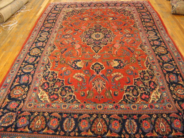 Ornate red and navy medallion rug displayed flat on polished hardwood flooring with another rolled rug nearby.