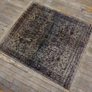 A distressed square rug with intricate dark purple and tan floral designs lies on gray wooden floorboards.