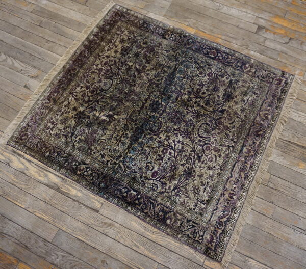 A distressed square rug with intricate dark purple and tan floral designs lies on gray wooden floorboards.