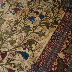Close-up of a beige rug with stylized animal heads, vines, and a deep burgundy patterned border on wood flooring.