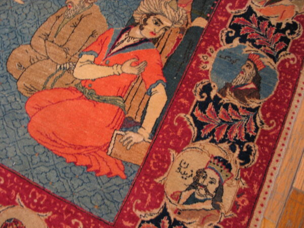 Low-pile rug detail showing figures in orange and tan on a blue field surrounded by a red border.