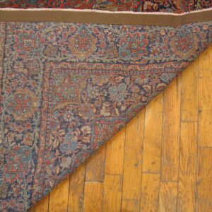 A dark red and navy patterned rug is folded back on wood flooring, revealing the reverse side and edge binding.