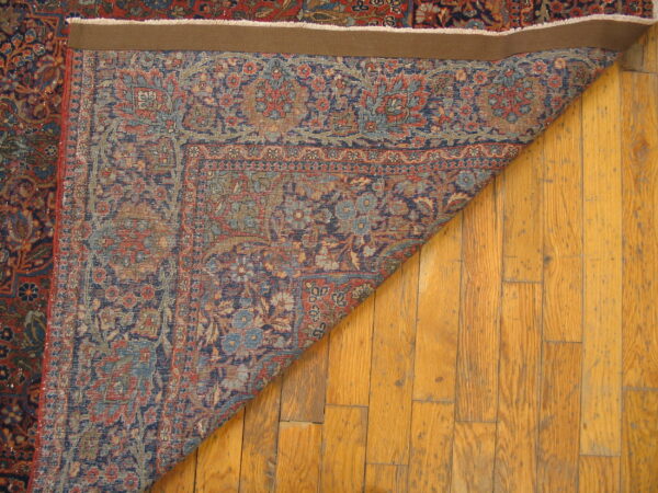 A dark red and navy patterned rug is folded back on wood flooring, revealing the reverse side and edge binding.