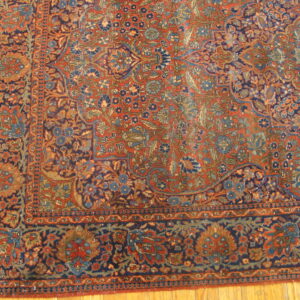 Corner of a rust and navy floral patterned rug with a detailed border on light hardwood flooring.