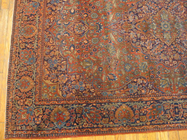 Corner of a rust and navy floral patterned rug with a detailed border on light hardwood flooring.