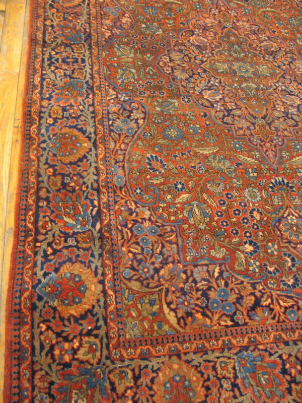 Close-up of a low-pile traditional rug with red, blue, and gold floral patterns over light wood flooring.