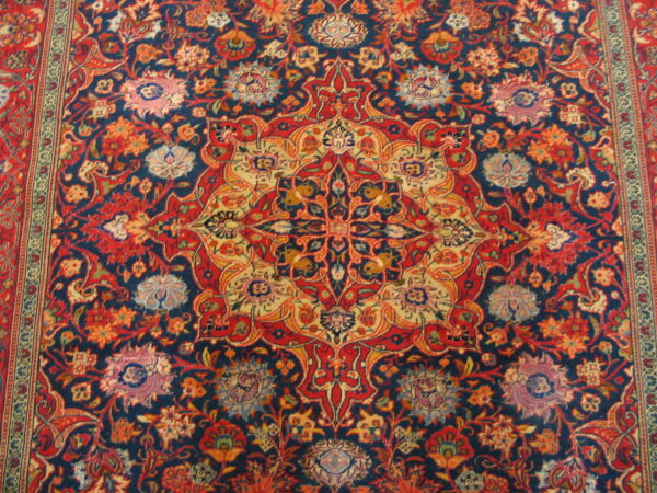 Intricate traditional rug pattern featuring a dense floral medallion motif in navy, red, and gold.