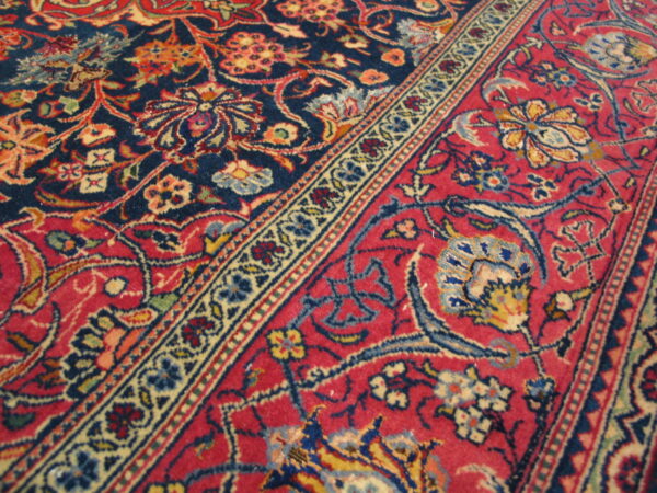 Angled close-up of a detailed rug pattern featuring red and navy blue fields separated by a cream floral border.