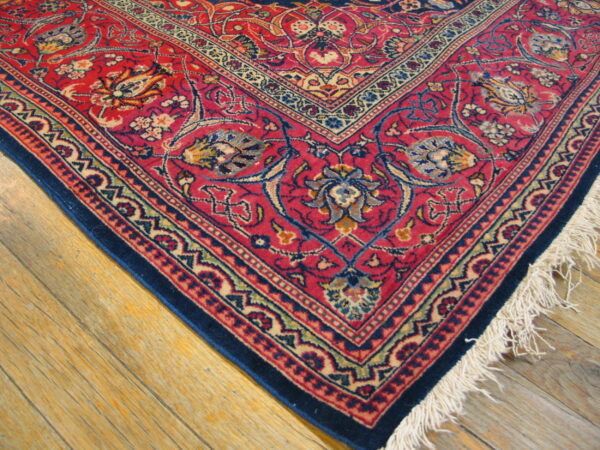 Intricate red and blue traditional rug corner showing floral motifs, white fringe, and distressed wood flooring.