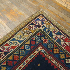 A corner of a geometric rug in navy, red, gold, and blue rests on light wood planks.