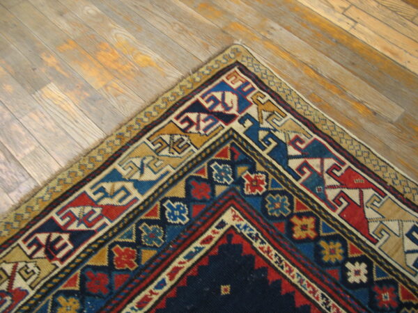 A corner of a geometric rug in navy, red, gold, and blue rests on light wood planks.