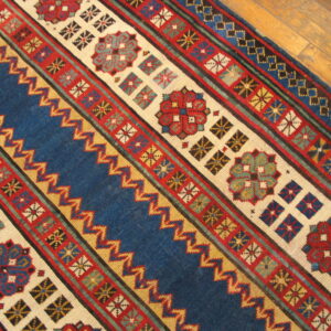 Blue, red, and cream geometric rug showing striped bands, medallions, and zigzag patterns on a wood floor.