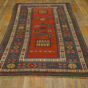 A long runner rug featuring a rust red field, teal border, and geometric motifs on distressed wood flooring.