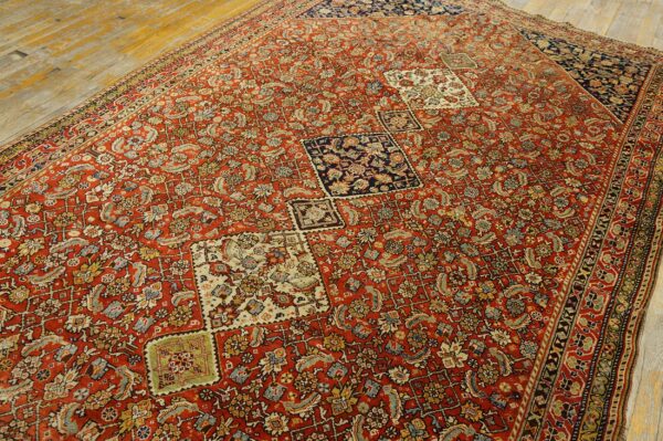 Low-pile red rug with dense traditional patterns, offset navy and cream squares, displayed on wood planks.
