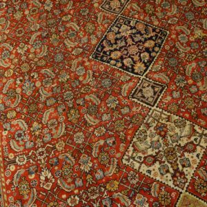 Intricate red rug featuring detailed floral patterns, a visible border, and contrasting squares on a wooden floor.