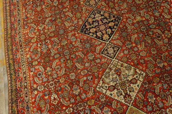 Intricate red rug featuring detailed floral patterns, a visible border, and contrasting squares on a wooden floor.