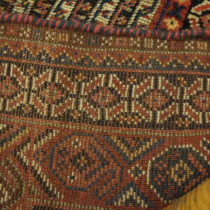 Tightly woven rug corner showing bands of geometric patterns in red, brown, blue, and black over wood planks.