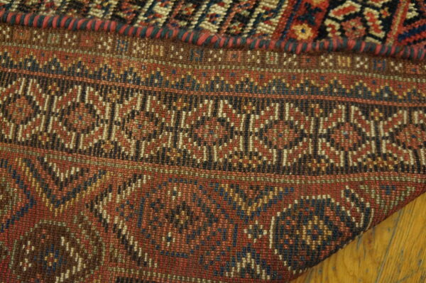 Tightly woven rug corner showing bands of geometric patterns in red, brown, blue, and black over wood planks.