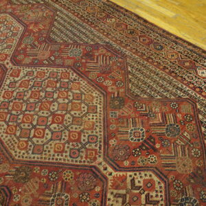 Geometric patterned rug in shades of red, cream, and black resting on light yellow wood plank flooring.