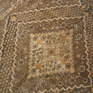 Brown and tan rug with dense traditional patterns, geometric borders, and a corner of wood flooring.