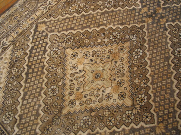 Brown and tan rug with dense traditional patterns, geometric borders, and a corner of wood flooring.