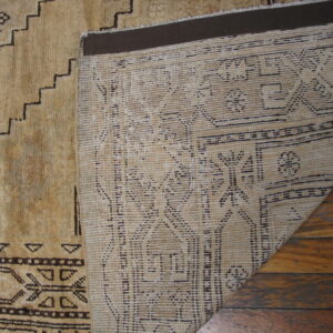 Geometric tan rug folded over, showing its dark patterned pile and woven backing on wood planks.