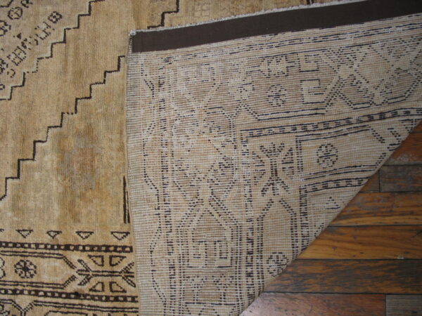 Geometric tan rug folded over, showing its dark patterned pile and woven backing on wood planks.