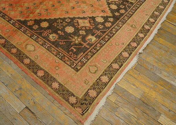 Close-up of a rug corner with coral, tan, and dark brown traditional patterns on distressed wooden floors.